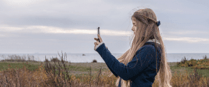 A young teenager with long blonde hair and wearing a blue sweater is standing in the grass near the shore of Lake Superior and is taking photographs with a smart phone she is holding in her hands.