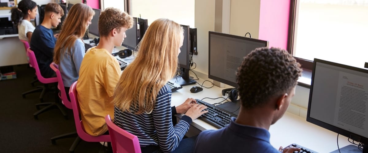 Several teenagers are sitting in a row on pink chairs each of them in front of a desktop computer.