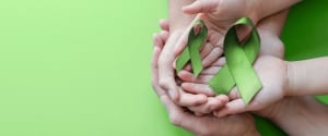 Two pair of hands rest inside one another holding two green ribbons upon a green background.